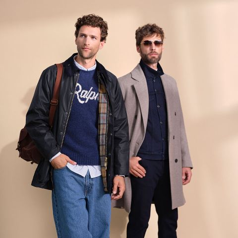 Due uomini posano di fronte a uno sfondo beige; uno indossa una giacca nera, jeans blu e un maglione blu navy con la scritta "Ralph", l'altro un cappotto grigio, pantaloni scuri e occhiali da sole.