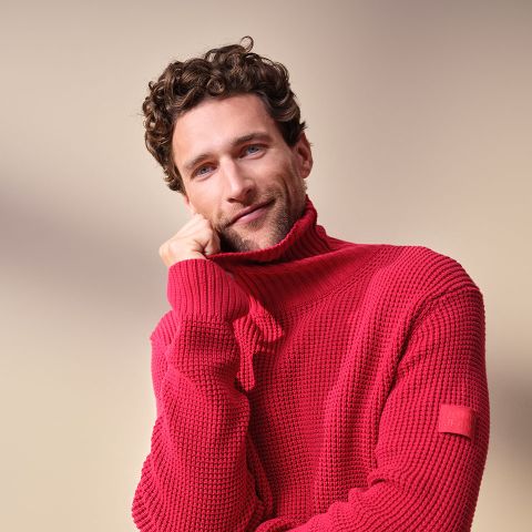 Un uomo con capelli castani ricci e occhi azzurri, che indossa un dolcevita rosso testurizzato, sorride dolcemente mentre appoggia il mento sulla mano davanti a uno sfondo beige.