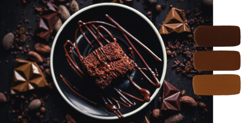 Un brownie al cioccolato con salsa al cioccolato su un piatto nero, circondato da scaglie di cioccolato e fave di cacao, con tre macchie di colore marrone sul lato destro dell'immagine.