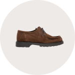 Ein brauner Wildlederschuh mit schwarzen Schnürsenkeln und einer dicken schwarzen Gummisohle, dargestellt im Profil auf einem hellbeigen runden Hintergrund.