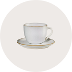 Eine weiße Teetasse aus Keramik mit Henkel steht auf einer passenden Untertasse. Beide haben einen dezenten Goldrand. Der Hintergrund ist ein leicht gräulicher Kreis.