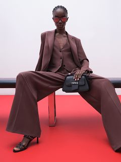 Una modella in un completo oversize marrone, pantaloni a gamba larga e tacchi neri è seduta su una panchina davanti a uno sfondo rosso e un muro bianco, indossa occhiali da sole e tiene in mano una borsa HUGO nera.