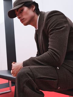Una persona in abito marrone e con un cappello abbinato è seduta su una panchina nera di fronte a uno sfondo rosso e bianco e guarda di lato con un'espressione pensierosa.