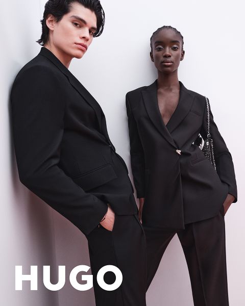 Due modelle in abiti neri posano con sicurezza davanti a uno sfondo chiaro e semplice. Il logo HUGO appare in grassetto bianco nell'angolo inferiore sinistro.