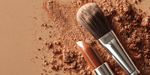 Un pennello da trucco e un rossetto color nude sono appoggiati su una superficie cosparsa di cipria marrone sciolta, il tutto in tonalità simili della terra.