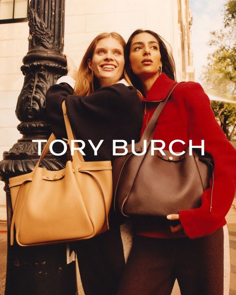 Due donne stanno all'aperto, abbracciate, sorridono, indossano cappotti eleganti e grandi borse di Tory Burch. La luce del sole mette in risalto i loro volti. Il testo "TORY BURCH" appare sopra l'immagine.