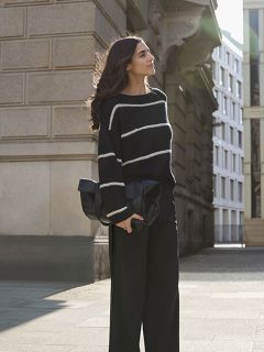 Una donna con i capelli lunghi è in piedi su un marciapiede in città. Indossa un maglione nero con strisce bianche e pantaloni neri a gamba larga e tiene in mano una grande pochette nera, mentre è illuminata dal sole da dietro.