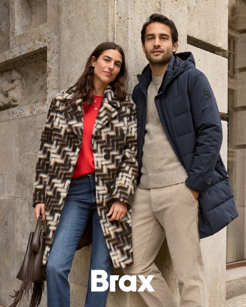 Una donna con un cappotto fantasia e un maglione rosso è in piedi accanto a un uomo con un cappotto invernale blu navy e pantaloni beige. Sono appoggiati a un muro di pietra all'aperto. La parola "Brax" appare nella parte inferiore dell'immagine.