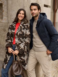 Una donna con un cappotto di pelliccia fantasia e un top rosso sorride accanto a un uomo con una giacca invernale blu navy e un maglione beige. Sono in piedi all'aperto vicino a un muro di pietra e la donna tiene in mano una borsa marrone.