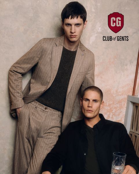 Due uomini posano per una foto di moda; uno indossa un abito gessato marrone chiaro e una camicia scura, mentre l'altro è seduto accanto a lui con una camicia nera. Lo sfondo è leggermente strutturato, con il logo del "CG Club of Gents" sopra di loro.