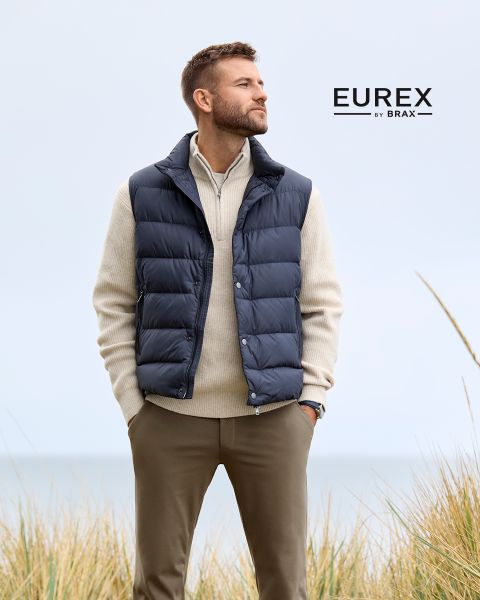 Un uomo è in piedi all'aperto vicino all'erba alta, indossa un maglione beige, un gilet imbottito blu navy e pantaloni marroni e guarda di lato. Il logo "EUREX by BRAX" appare sul lato destro. Il cielo è nuvoloso.
