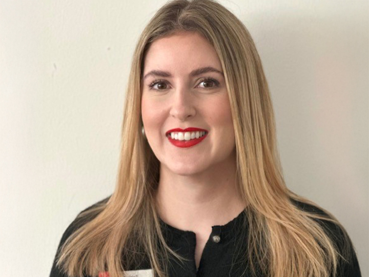 Donna con lunghi capelli biondi e rossetto rosso, che indossa un top nero, sorride davanti a uno sfondo chiaro e semplice.