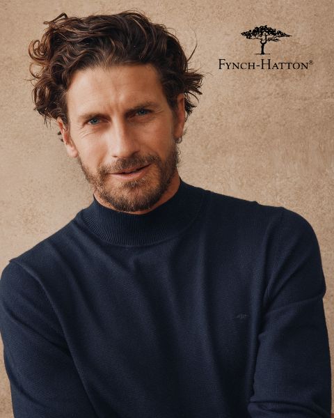 Un uomo con capelli castani ondulati e barba indossa un maglione blu navy e si trova di fronte a una parete beige strutturata. Il logo e il nome di Fynch-Hatton appaiono nell'angolo in alto a destra.