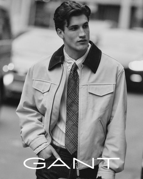 Un uomo che indossa una camicia con colletto, una cravatta e una giacca chiara esce all'aperto con le mani in tasca. L'immagine è in bianco e nero, con la scritta "GANT" in basso.