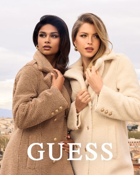 Due donne sono in piedi vicine all'aperto, indossando cappotti strutturati, una in marrone chiaro e una in color crema. Entrambe tengono stretto il colletto e guardano avanti. La parola "GUESS" è scritta in grassetto bianco in basso.