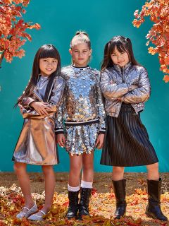 Tre giovani ragazze posano con sicurezza tra le foglie autunnali, indossando abiti metallici lucenti. Due ragazze indossano giacche argentate e la ragazza al centro indossa un top e una gonna di paillettes. Uno sfondo verde acqua contrasta con il fogliame autunnale arancione.