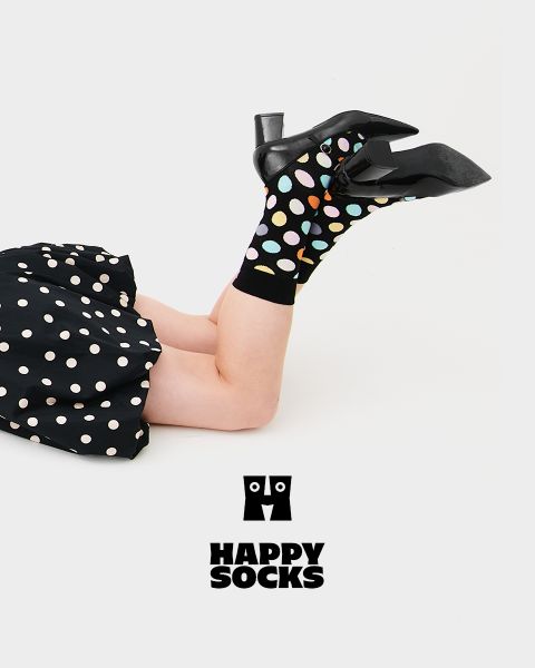 Una persona con scarpe col tacco nere e calzini neri con pois colorati è sdraiata su un fianco, con le gambe in alto, su uno sfondo bianco. Indossa una gonna nera a pois. Sotto appare il logo Happy Socks.