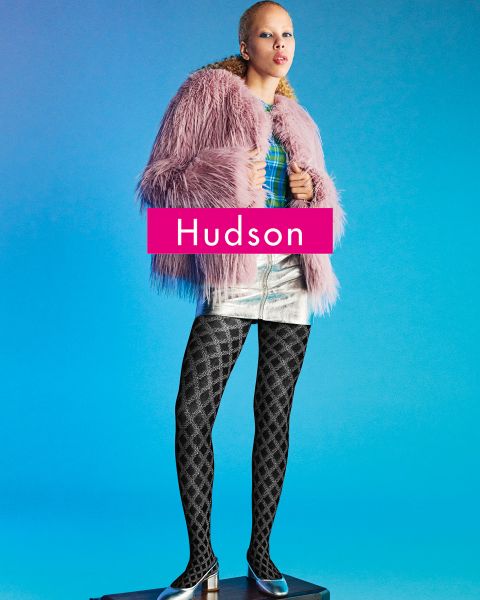 Una persona è in piedi su un piedistallo davanti a uno sfondo blu, indossa un cappotto di pelliccia sintetica rosa, un top a quadri, una gonna metallizzata argentata, collant neri fantasia e scarpe a punta. La parola "Hudson" appare in grassetto sull'immagine.