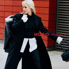 Una donna in cappotto nero e camicia bianca posa accanto alla scritta "HUGO RED MEANS GO".