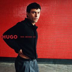 Un uomo con un cardigan nero e pantaloni grigi si trova davanti a una parete di piastrelle rosse con la scritta "HUGO RED MEANS GO".