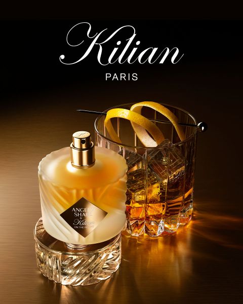 Un flacone del profumo Kilian Paris Angels' Share è accanto a un bicchiere di whisky con ghiaccio e guarnizione, entrambi su una superficie marrone riflettente con illuminazione soffusa. In alto è visibile il logo Kilian Paris.