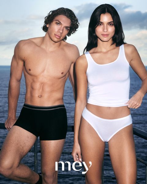 Un uomo in boxer neri e una donna in canottiera bianca e biancheria intima sono in piedi uno accanto all'altra all'aperto vicino a una ringhiera, con il mare e il cielo nuvoloso sullo sfondo. La parola "mey" è in basso.