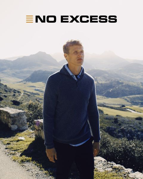 Un uomo con un maglione blu è in piedi all'aperto in una giornata di sole, con montagne e valli sullo sfondo. Le parole "NO EXCESS" appaiono in grassetto nella parte superiore dell'immagine.