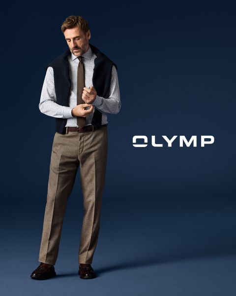 Un uomo in pantaloni da abito, camicia a quadri e cravatta con un maglione drappeggiato sulle spalle è in piedi davanti a uno sfondo blu scuro, accanto al quale si legge la parola "OLYMP".