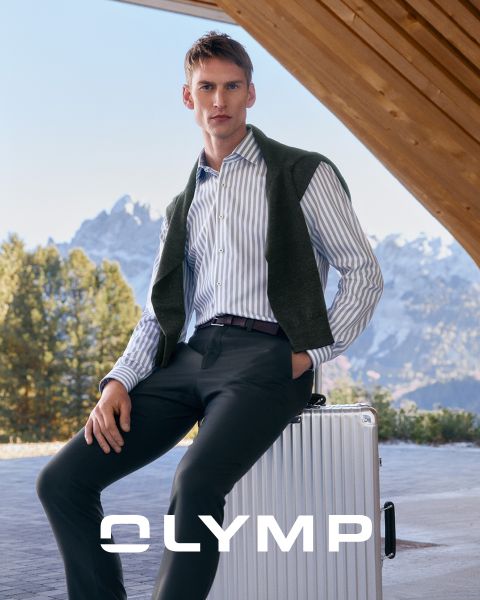 Un uomo con una camicia a righe e pantaloni scuri è seduto su una valigia argentata all'aperto, con un maglione appoggiato sulle spalle. Sullo sfondo si vedono montagne innevate e alberi. In basso c'è la parola "OLYMP".