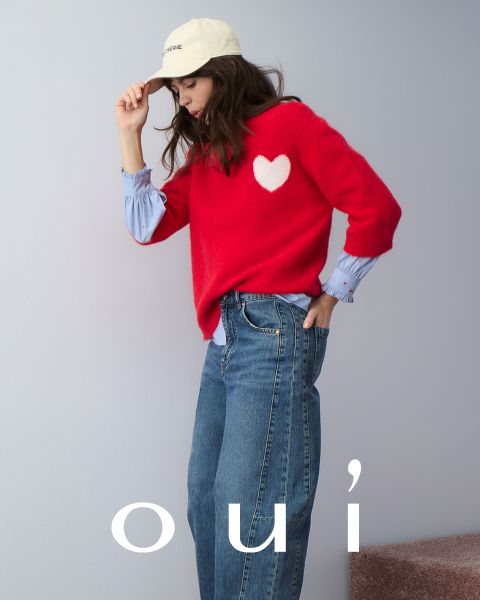 Una donna che indossa un maglione rosso con un cuore bianco, jeans blu e un cappello bianco posa davanti a un muro chiaro. Nella parte inferiore dell'immagine c'è la parola "oui" in lettere bianche.