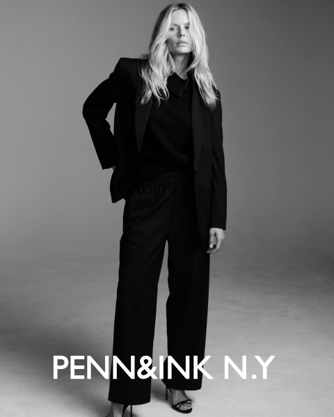 Una donna con lunghi capelli ondulati indossa un abito scuro e sandali con tacco alto, e posa con sicurezza in uno studio con sfondo monocolore. Il testo in basso recita "PENN&INK N.Y."