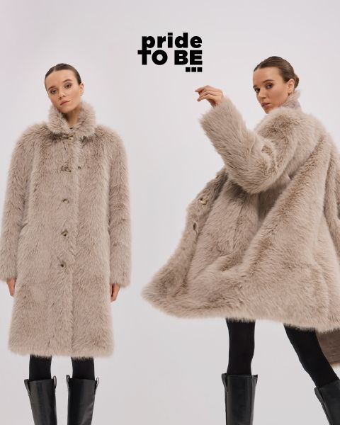 Due donne con lunghi cappotti di pelliccia sintetica beige e stivali neri posano davanti a uno sfondo semplice. Sopra di loro c'è il testo "pride TO BE". Una donna è in piedi con la faccia rivolta in avanti, l'altra si gira di lato con un braccio alzato.