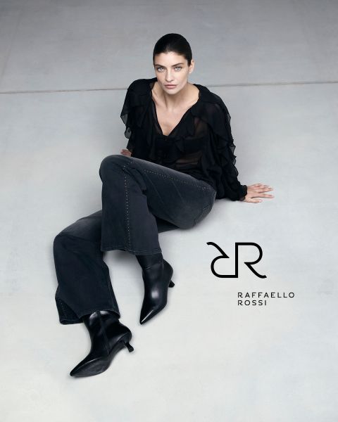 Una donna con i capelli scuri è seduta su un pavimento grigio chiaro e indossa una blusa nera con volant, jeans scuri e stivali neri a punta. Accanto a lei si vede il logo di Raffaello Rossi.