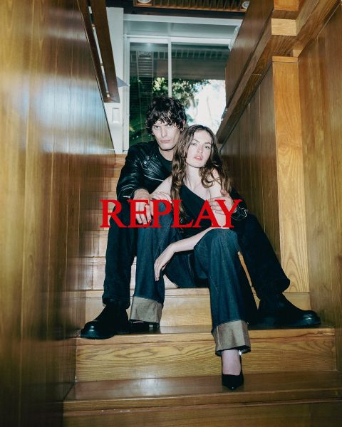Un uomo e una donna siedono su una scala di legno in un interno, vestiti con abiti scuri ed eleganti. La parola "REPLAY" è sovraimpressa in modo prominente al centro dell'immagine in lettere maiuscole rosse.
