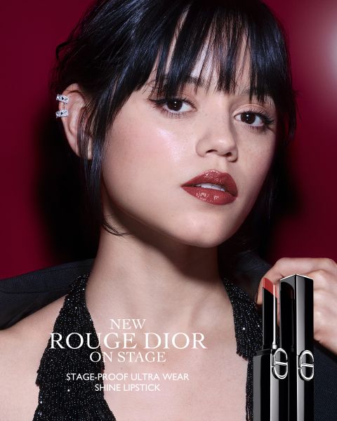 Una donna con un rossetto rosso scuro lucido posa davanti a uno sfondo rosso intenso, indossando un top nero luccicante all'americana e degli ear cuff. Tiene in mano un tubetto di Rouge Dior Shine Lipstick. Il testo pubblicizza il nuovo rossetto ultra-wear a prova di palcoscenico.