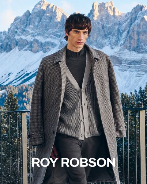 Un uomo è in piedi all'aperto di fronte a montagne innevate e indossa un cappotto pied-de-poule grigio, un maglione grigio e pantaloni grigi. Il testo "ROY ROBSON" appare nella parte inferiore dell'immagine.