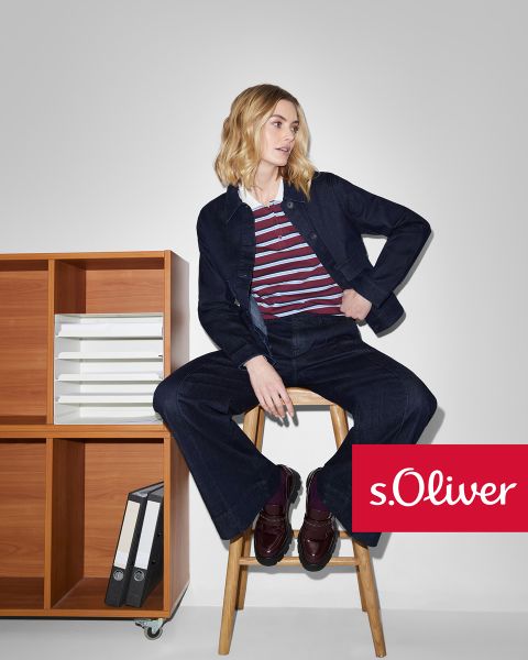 Una donna con giacca e pantaloni di jeans scuri e una maglietta a righe è seduta su uno sgabello di legno accanto a uno scaffale di legno con dei documenti. Il logo s.Oliver appare nell'angolo in basso a destra.