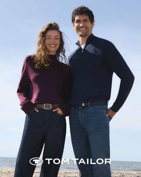Una donna sorridente con un maglione color castagna e un uomo con un maglione blu navy sono in piedi fianco a fianco su una spiaggia. Entrambi indossano jeans. Il cielo è limpido e blu. Il logo "Tom Tailor" appare in basso.
