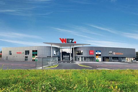 Un moderno centro commerciale con un grande cartello "WEZ" sul tetto, una facciata grigia, diverse insegne di negozi e un cielo azzurro limpido sopra.