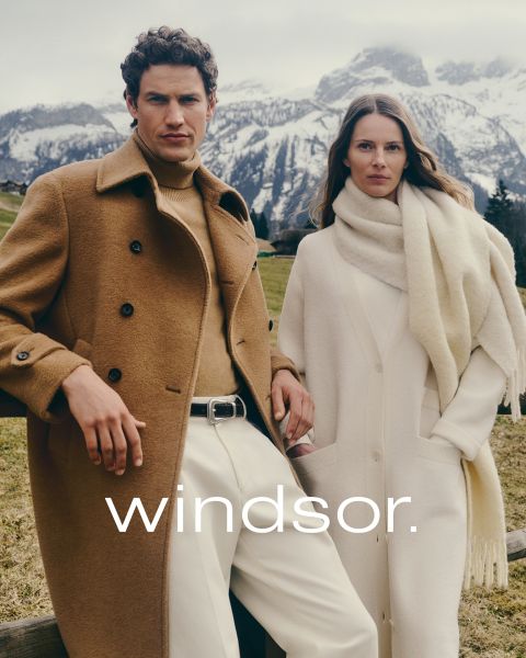 Un uomo con un cappotto marrone e una donna con un cappotto e una sciarpa color crema sono in piedi all'aperto con le montagne sullo sfondo. La parola "windsor." è visibile nella parte inferiore dell'immagine.
