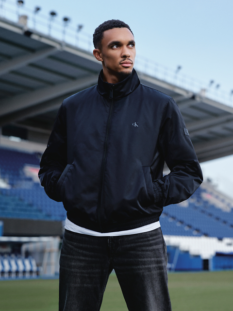 Un uomo con una giacca nera e jeans è in piedi in uno stadio e guarda di lato con le mani in tasca. Sullo sfondo si vedono i posti a sedere dello stadio e un grande edificio.