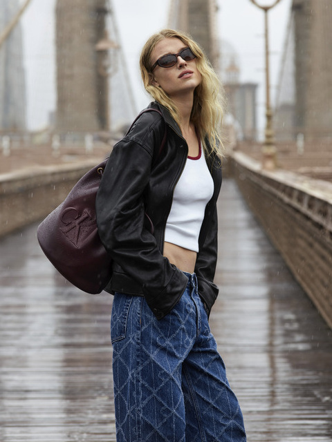 Una donna con occhiali da sole, giacca nera, top corto bianco e ampi jeans blu è in piedi su un ponte piovoso e porta una grande borsa marrone castagna a tracolla. Il ponte e le lampade sono sfocati sullo sfondo.