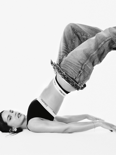 Foto in bianco e nero di una donna che indossa un reggiseno sportivo Calvin Klein e jeans, sdraiata sulla schiena con le gambe piegate sopra la testa, le braccia distese sul pavimento e lo sguardo rivolto alla fotocamera.