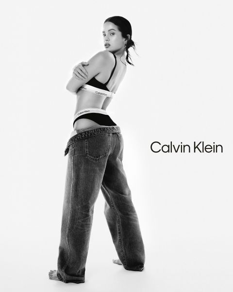 Una donna posa in biancheria intima Calvin Klein e jeans larghi, guardando oltre la sua spalla. Lo sfondo è bianco uniforme con il logo Calvin Klein in caratteri neri sul lato destro. L'immagine è in bianco e nero.