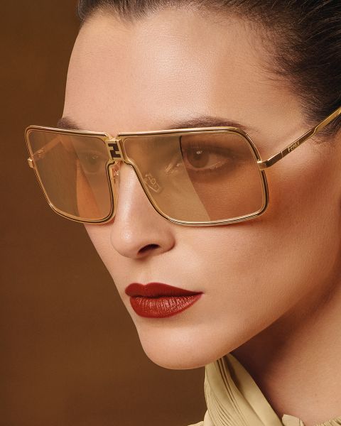 Primo piano di una persona che indossa grandi occhiali da sole quadrati con montatura dorata e lenti marroni. Ha la pelle liscia, i capelli tirati indietro, indossa un rossetto rosso acceso e un top chiaro su uno sfondo marrone.