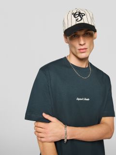 Un giovane uomo che indossa una maglietta nera con la scritta "Original Studio", una collana d'argento, un braccialetto d'argento e un berretto da baseball a righe color crema e nero, è in piedi davanti a uno sfondo semplice grigio chiaro.