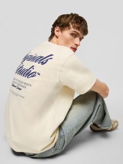 Un giovane uomo con capelli castani corti, che indossa una maglietta color crema con una stampa blu navy sul retro, jeans azzurri e scarpe da ginnastica beige, è seduto sul pavimento e guarda la telecamera sopra la spalla.