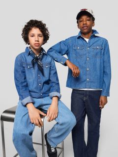 Due ragazzi in jeans e camicie di jeans posano insieme. Uno è seduto su uno sgabello con le mani in grembo e una sciarpa al collo, mentre l'altro ha un braccio sullo sgabello e indossa un berretto.