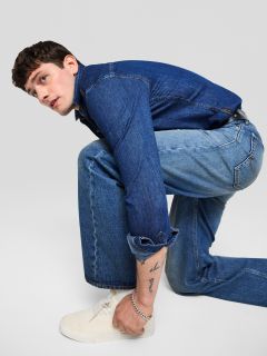 Un giovane uomo in giacca di jeans e jeans è inginocchiato e guarda di lato. Indossa scarpe da ginnastica bianche e ha un tatuaggio visibile sull'avambraccio. Lo sfondo è grigio chiaro uniforme.