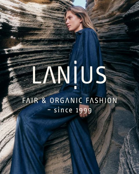 Una donna con un lungo abito blu scuro è seduta tra formazioni rocciose stratificate. Il testo sovrapposto recita: "LANIUS FAIR & ORGANIC FASHION - dal 1999.